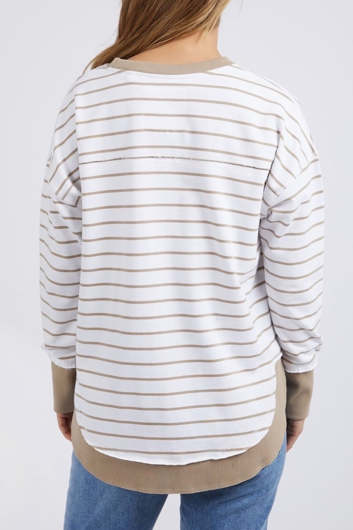 The back view of an individual wearing the Foxwood Simplified Stripe Crew is showcased, highlighting its thin beige yarn dye stripes, relaxed fit, and curved hemline. This chic piece from Foxwood Clothing is complemented beautifully with blue jeans.