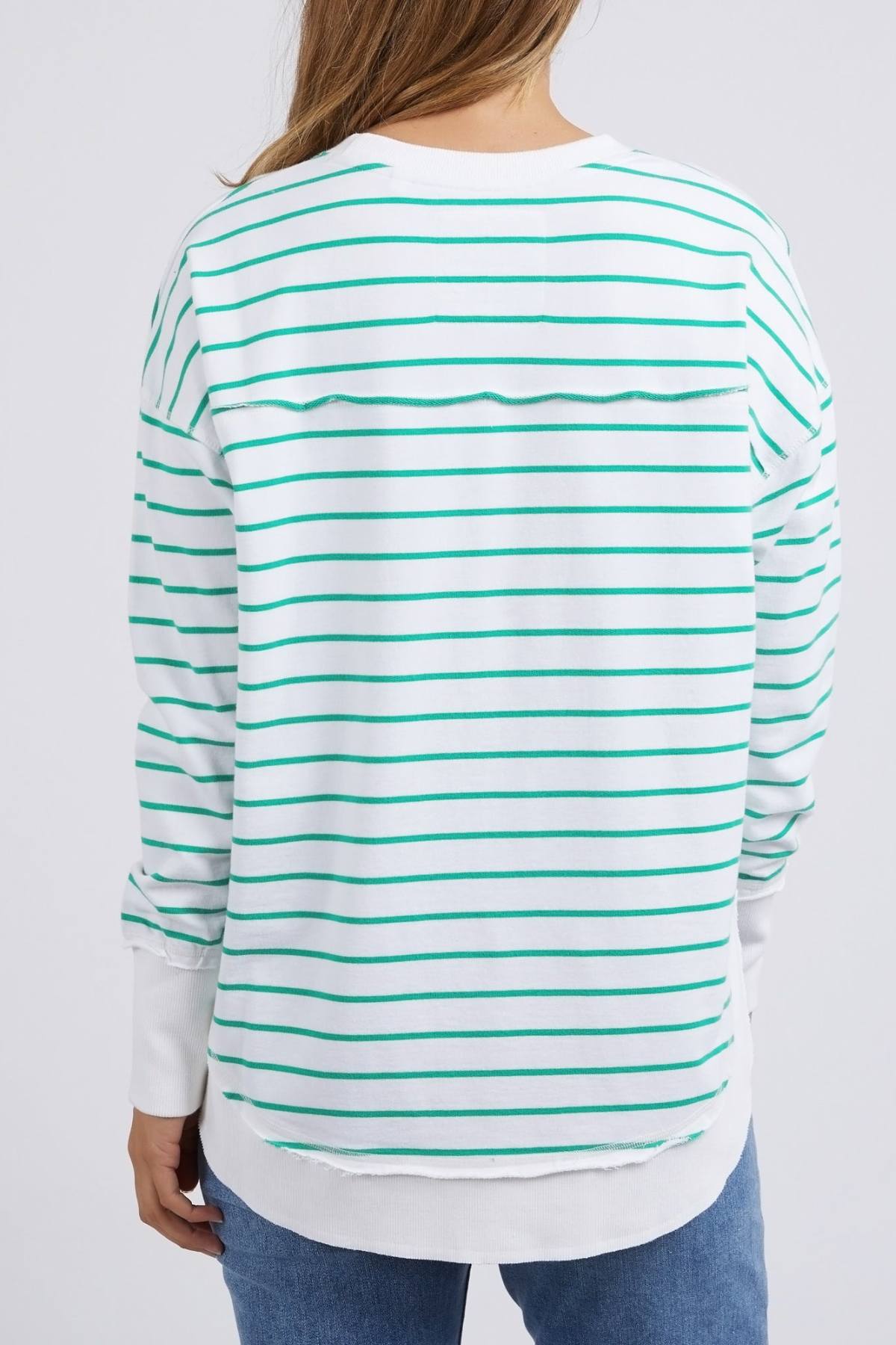 A person is seen from the back wearing the Foxwood Simplified Stripe Crew, a relaxed-fit sweater made of 100% cotton. It features white and green horizontal stripes, long sleeves, and a straight hem, paired with blue jeans. Brown hair flows over their shoulders against a plain white background.