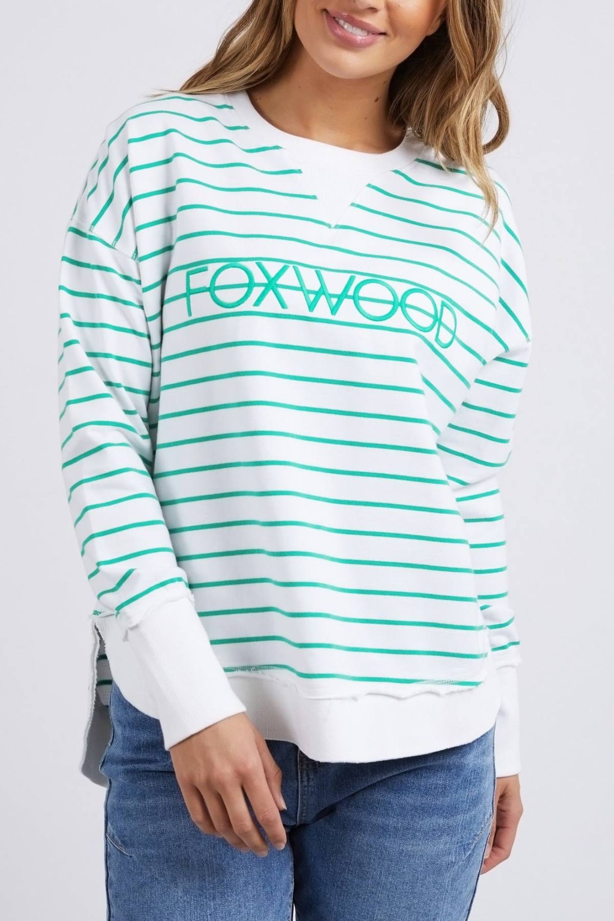 A person is wearing a Foxwood Simplified Stripe Crew sweatshirt, featuring green stripes and "FOXWOOD" printed on the front. Made from 100% cotton, it offers a relaxed fit. They pair it with blue jeans, and their long hair completes the look against a plain white background.
