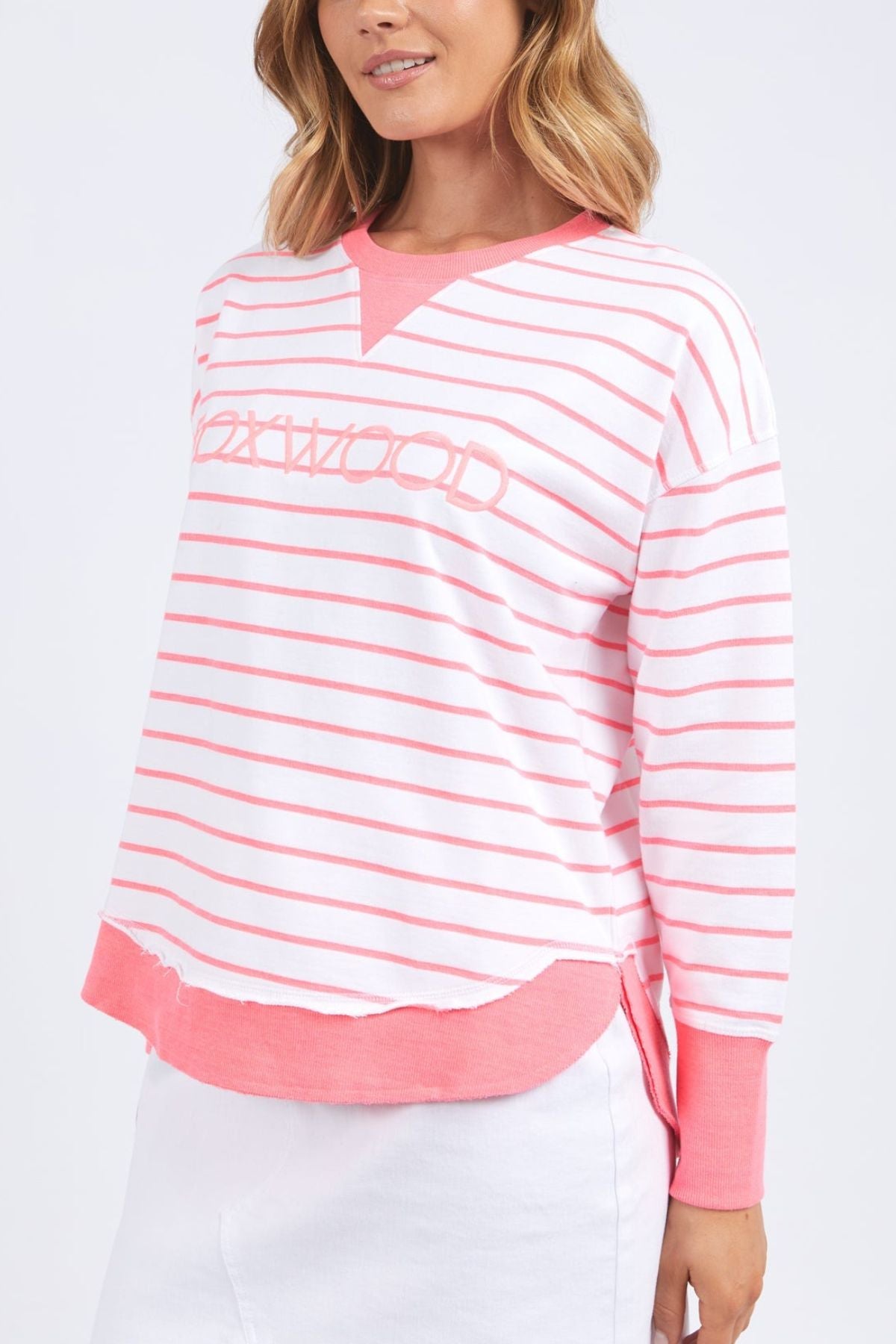 A person is dressed in the Foxwood Simplified Stripe Crew, a pink and white striped long-sleeve shirt with "Foxwood" on it. This shirt, perfect for layering, has a pink neckline and cuffs. They have shoulder-length wavy hair and are paired with a white skirt.