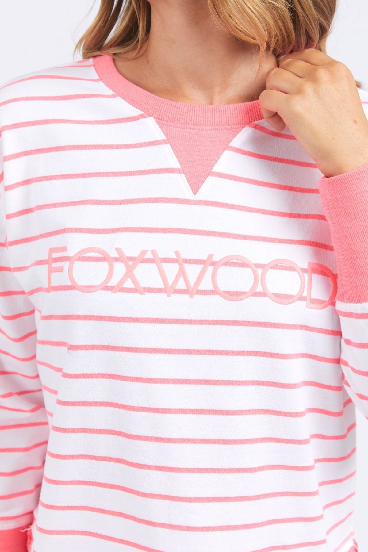 A person is seen in the Foxwood Simplified Stripe Crew, a stylish white and pink sweatshirt by Foxwood Clothing, featuring "FOXWOOD" boldly printed on the front. It's an ideal layering option with its cozy pink cuffs and collar.