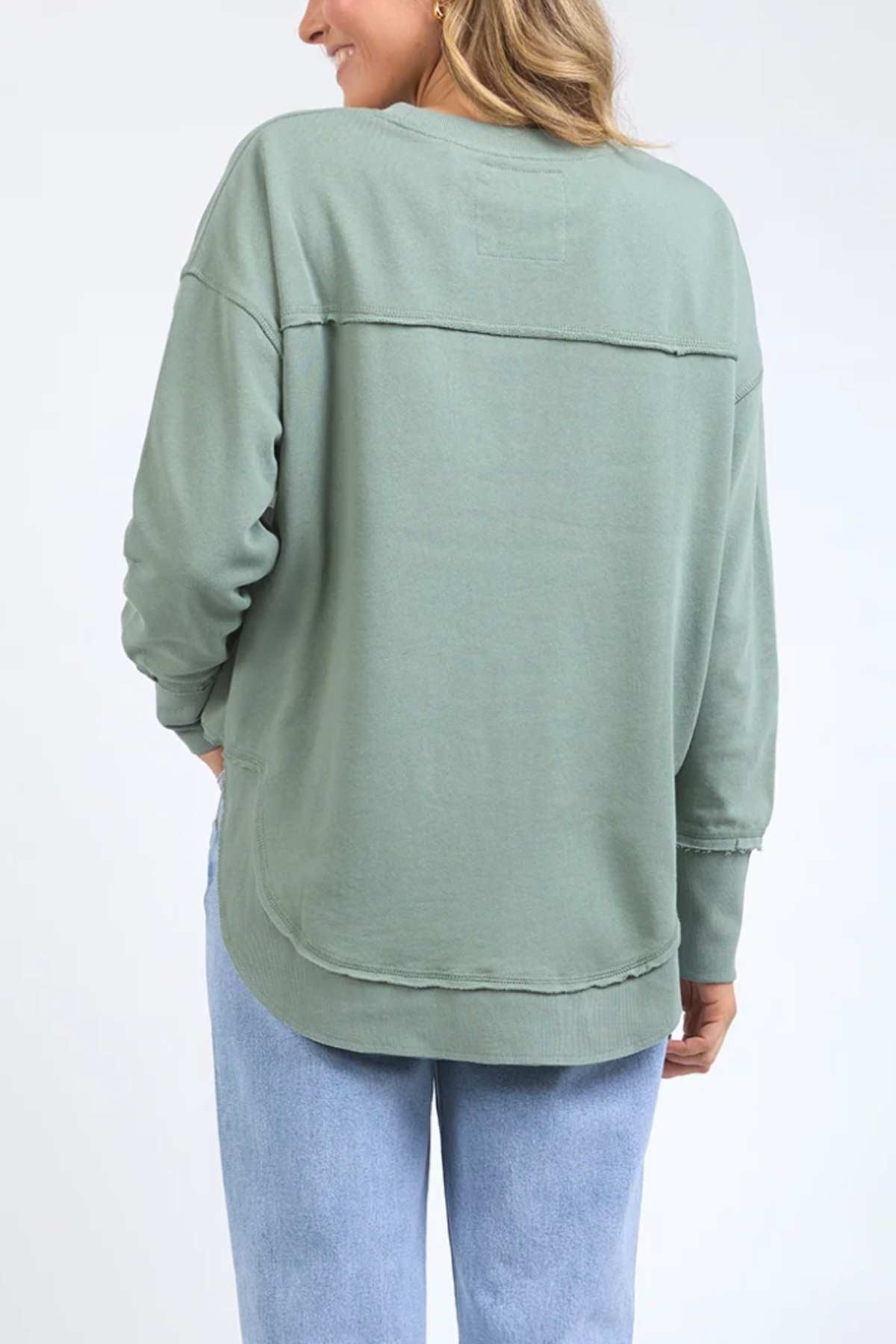 A person in a light green Foxwood Simplified Tonal Crew sweatshirt with raw edge detailing and light blue jeans stands with their back turned. The cotton fleece outfit creates a casual look against a plain white background.