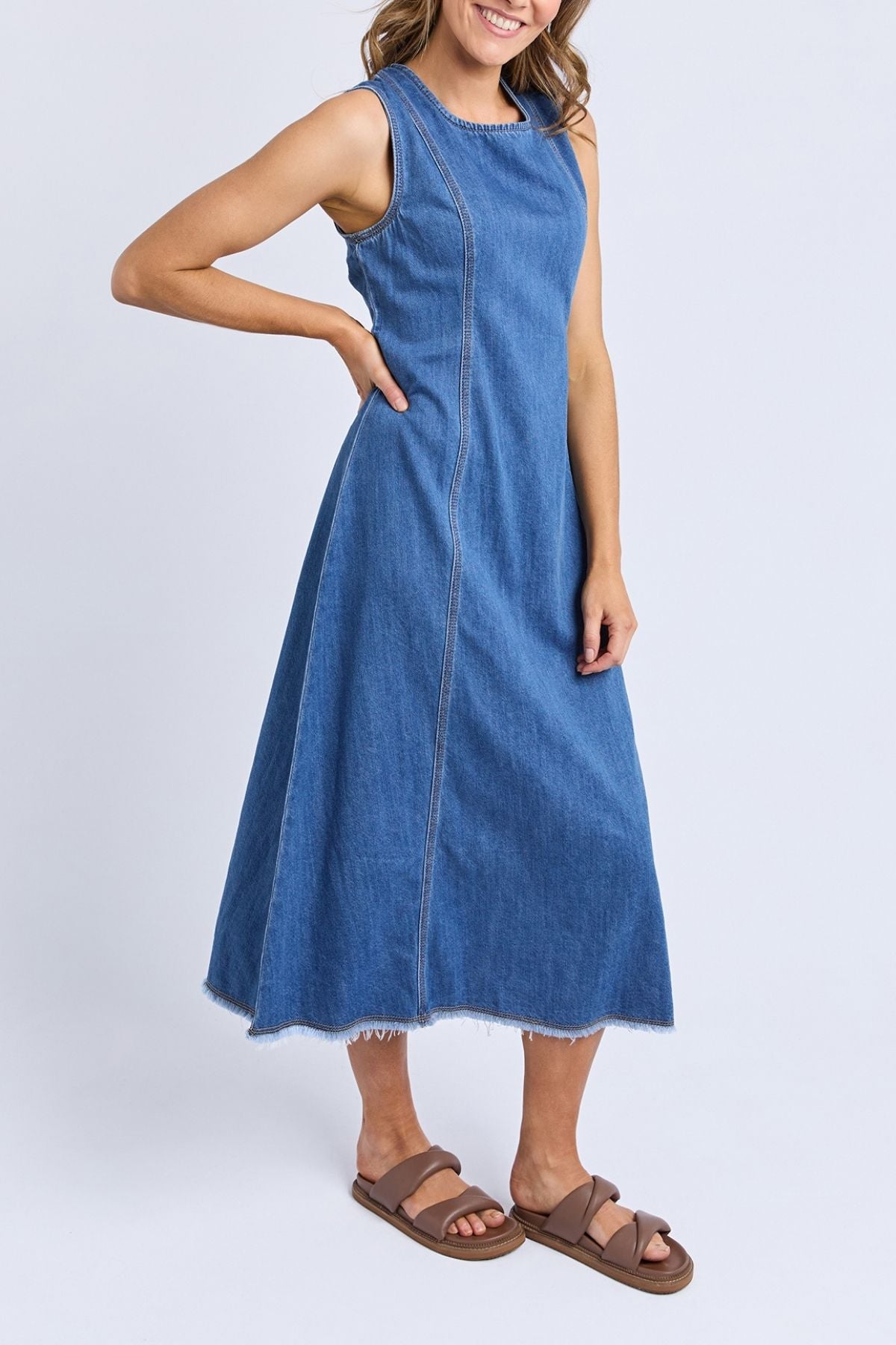 A woman stands against a light background, smiling with one hand on her hip, wearing the Foxwood Solene Denim Dress featuring an A-line silhouette, exposed seams, and raw hem detailing, paired with brown sandals.