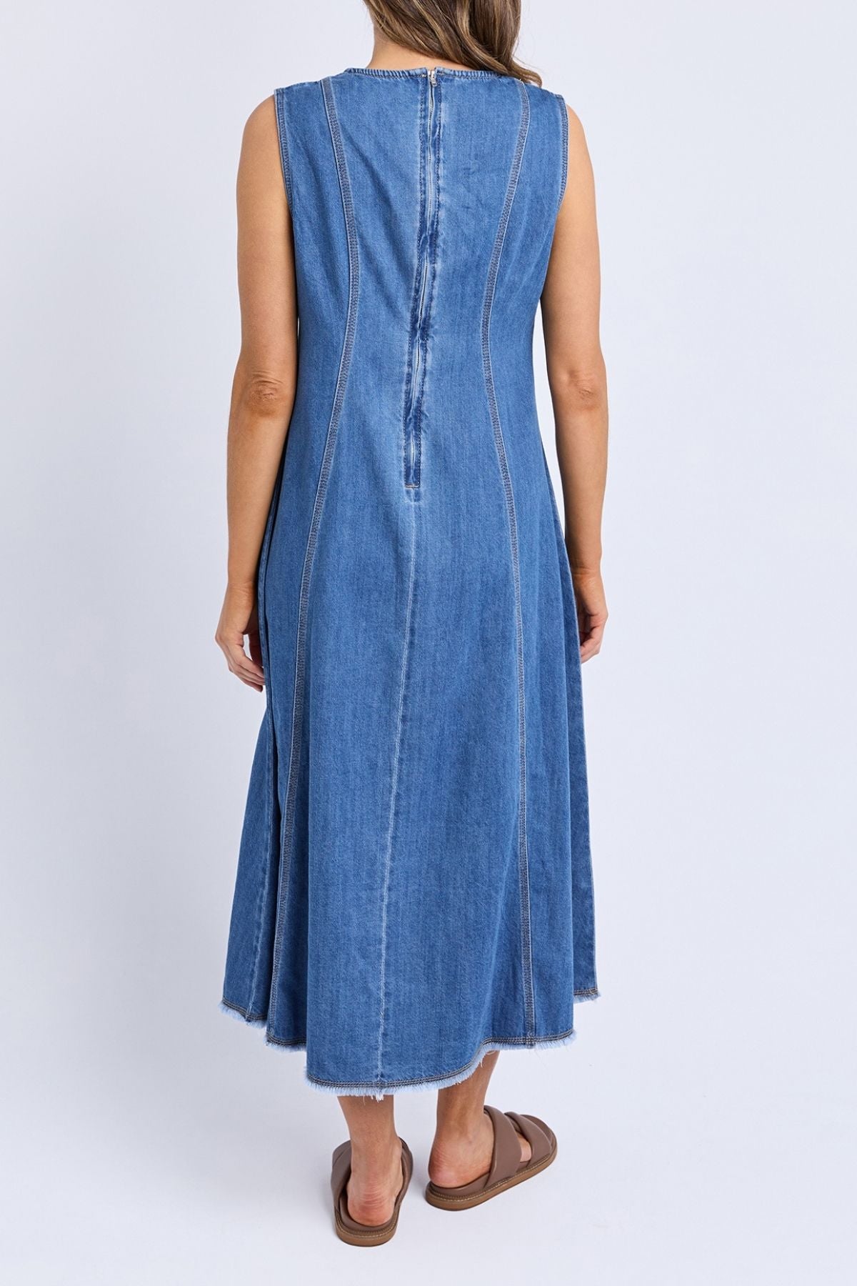 A woman faces away, wearing the Foxwood Solene Denim Dress—a sleeveless, A-line blue denim piece with visible seams and a raw hem—paired with brown slide sandals, set against a plain white background.