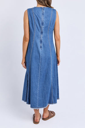 A woman faces away, wearing the Foxwood Solene Denim Dress—a sleeveless, A-line blue denim piece with visible seams and a raw hem—paired with brown slide sandals, set against a plain white background.