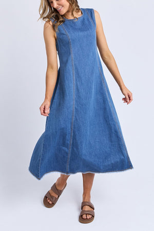 A woman in the Foxwood Solene Denim Dress—sleeveless, A-line, with raw hems and visible seams—smiles as she gently sways her skirt, paired with brown sandals against a plain light background.