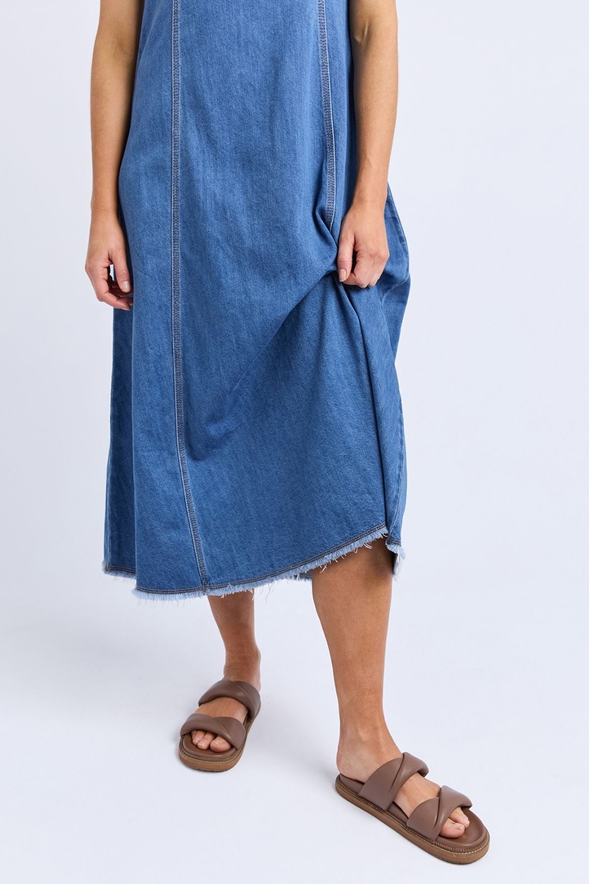 A person wears the Foxwood Solene Denim Dress—a sleeveless blue denim dress with raw hem and A-line silhouette—holding one side. They pair it with brown slide sandals against a plain white background.