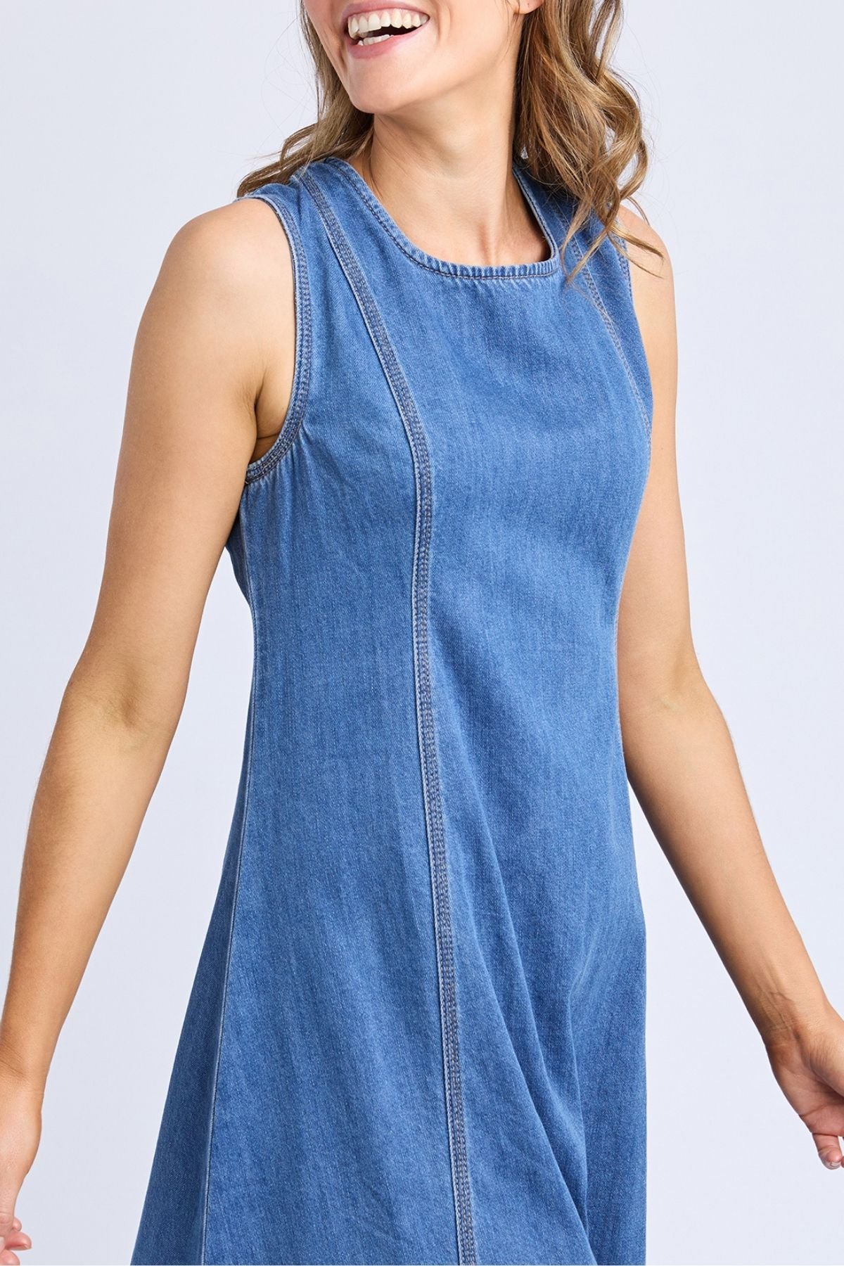 A woman with loose, wavy hair smiles while wearing the Foxwood Solene Denim Dress, a sleeveless A-line blue denim style. Only her upper body is visible against a plain, light background.