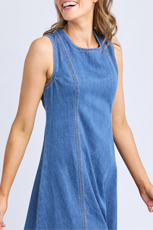 A woman with loose, wavy hair smiles while wearing the Foxwood Solene Denim Dress, a sleeveless A-line blue denim style. Only her upper body is visible against a plain, light background.