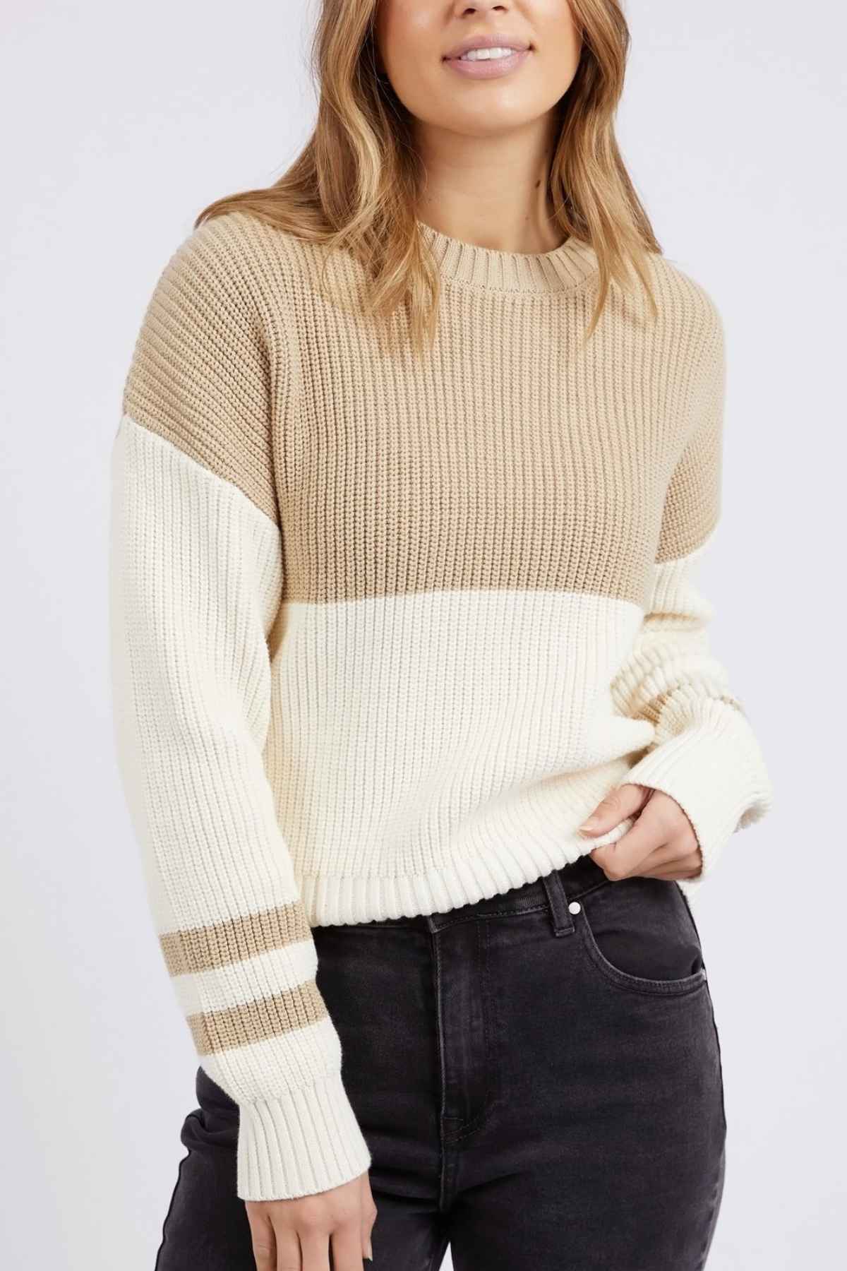 A person with long hair is wearing the Foxwood Sophie Knit Crew, a relaxed-fit beige and white color-block sweater featuring striped sleeves, paired with dark jeans. They are standing against a plain background with one hand partially in their jeans pocket.