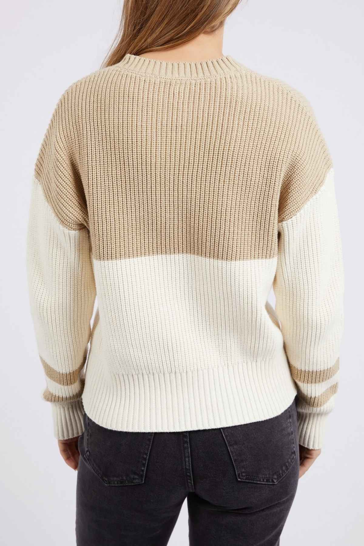  A person with long hair is seen from behind, wearing a relaxed-fit Foxwood Sophie Knit Crew in beige, tan, and white with ribbed textures. The person is also wearing dark jeans. The background is plain and light-colored.