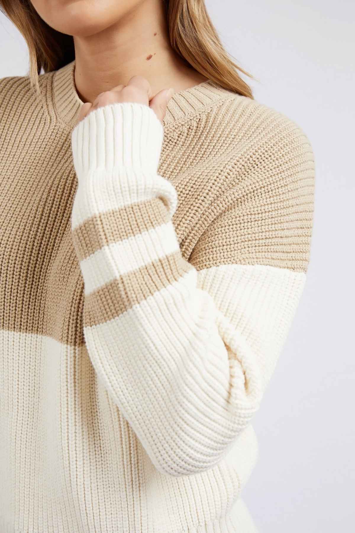 A person with shoulder-length hair is wearing a relaxed-fit Foxwood Sophie Knit Crew, showcasing its ribbed texture and stylish color blocking in shades of beige, white, and taupe. The sweater features horizontal stripes. The person's face is partially out of the frame as they hold up the sleeve of the sweater.