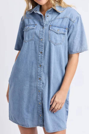 A woman wearing the Foxwood Sorcha Denim Dress—a light blue, short-sleeve denim shirt dress with front buttons and two chest pockets—stands with one hand gently placed in her pocket.