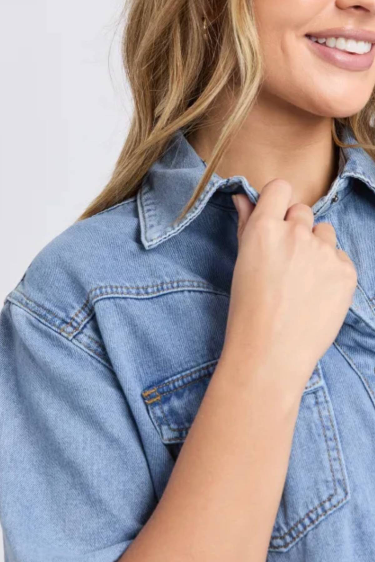 A woman with long blonde hair smiles, holding the collar of her light blue Foxwood Sorcha Denim Dress. Only the lower half of her face and upper torso are visible.