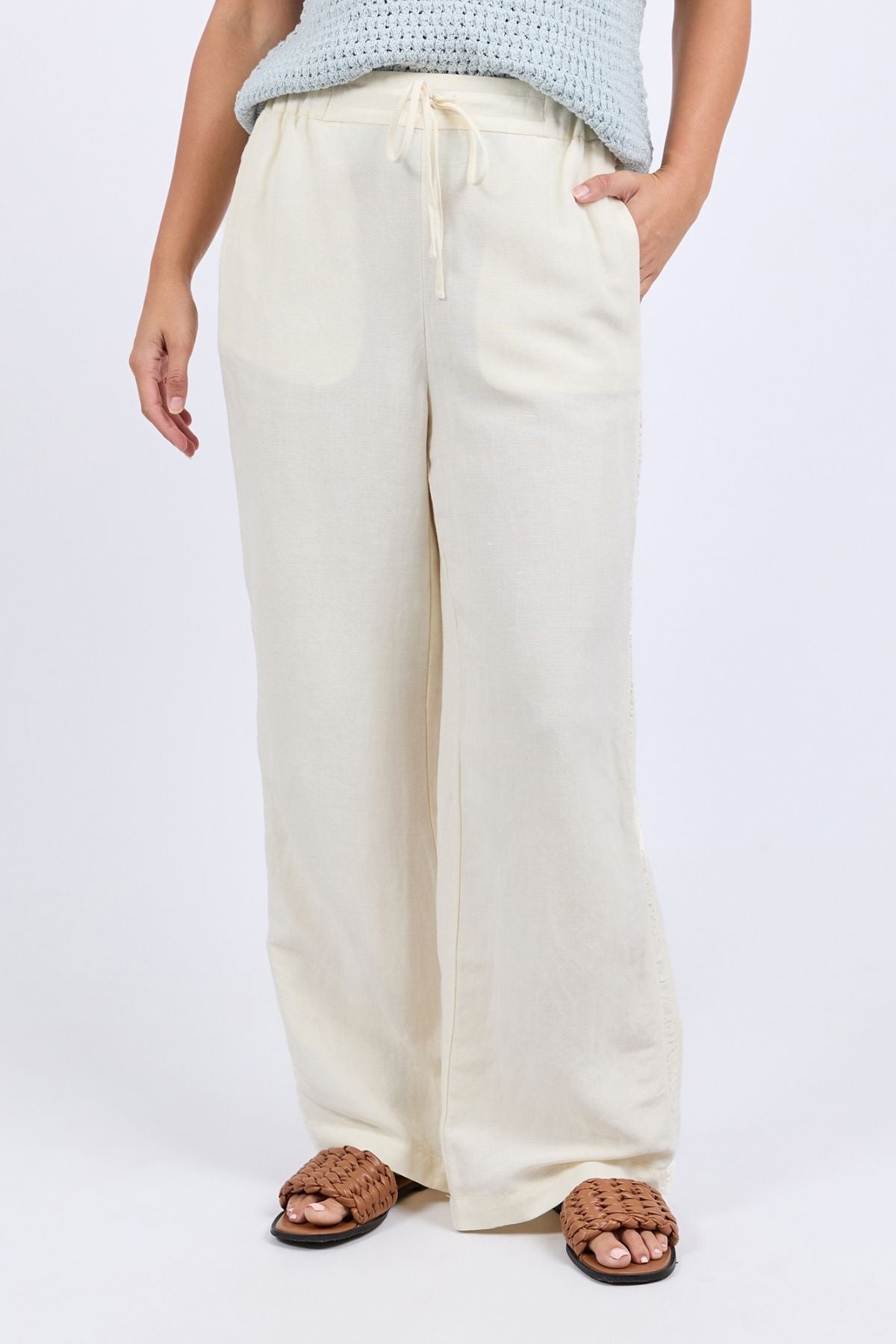 A person wears the Foxwood South Pant—women's cream linen pants with an elastic waist—styled with a light blue knit top and brown woven sandals, standing with one hand in their pocket against a plain backdrop.