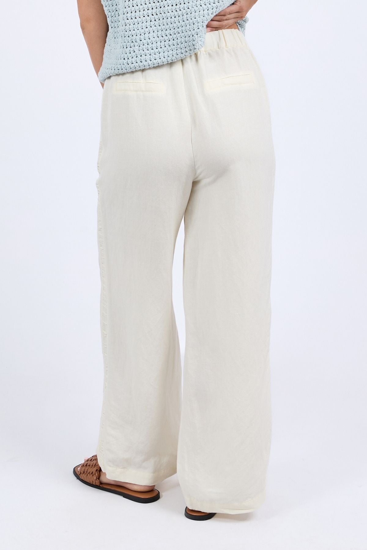Woman wearing the Foxwood South Pant—a loose, cream-colored linen pant with an easy elastic waist—paired with a light blue crochet top and brown woven sandals, shown from the back for a relaxed vibe.