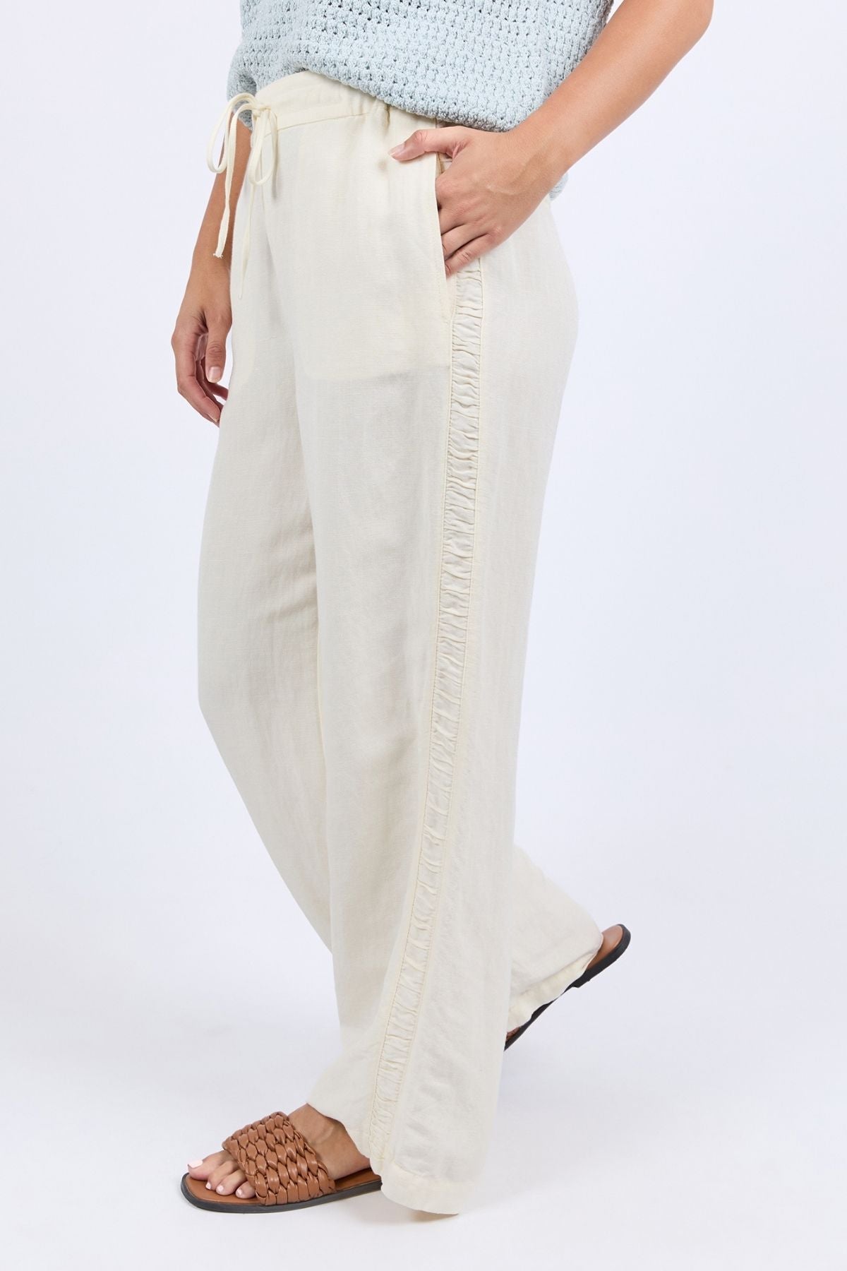 A woman wears the Foxwood South Pant—light beige, wide-leg linen pants with a drawstring waist and ruffled trim—paired with brown woven sandals and a light blue knit top for effortless style and comfort.