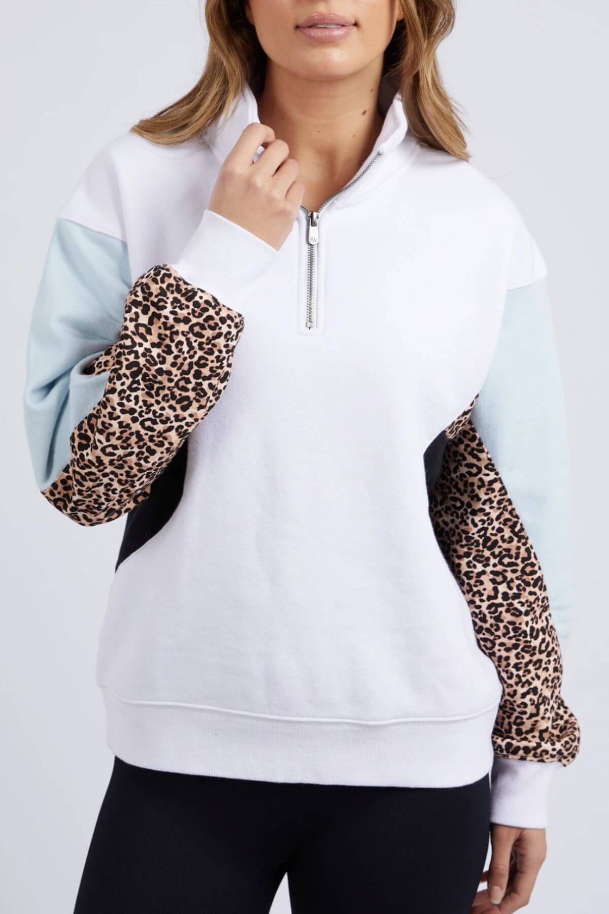 A woman wears the Foxwood Splice Zip Crew, a white cotton quarter-zip pullover with light blue and leopard print color-block sleeves and a relaxed fit, paired with black pants.