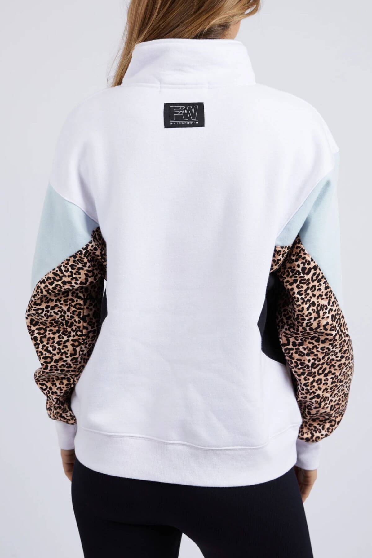 A woman is seen from behind wearing the Foxwood Splice Zip Crew, a relaxed fit white cotton sweatshirt with a black collar label, pale blue shoulder panels, and leopard print sleeves. She pairs it with black leggings.