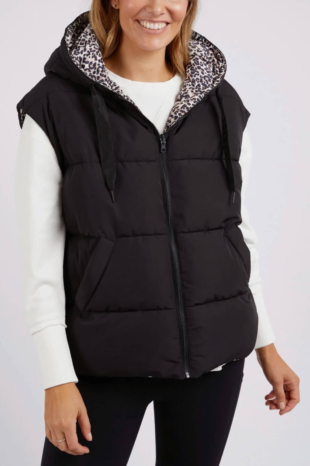 A person models the Foxwood Sports Leopard Vest, featuring a black puffer design with a leopard print hood lining and adjustable toggles, paired with a white long-sleeve top against a plain backdrop. The vest's front zipper and pocket details add stylish functionality.