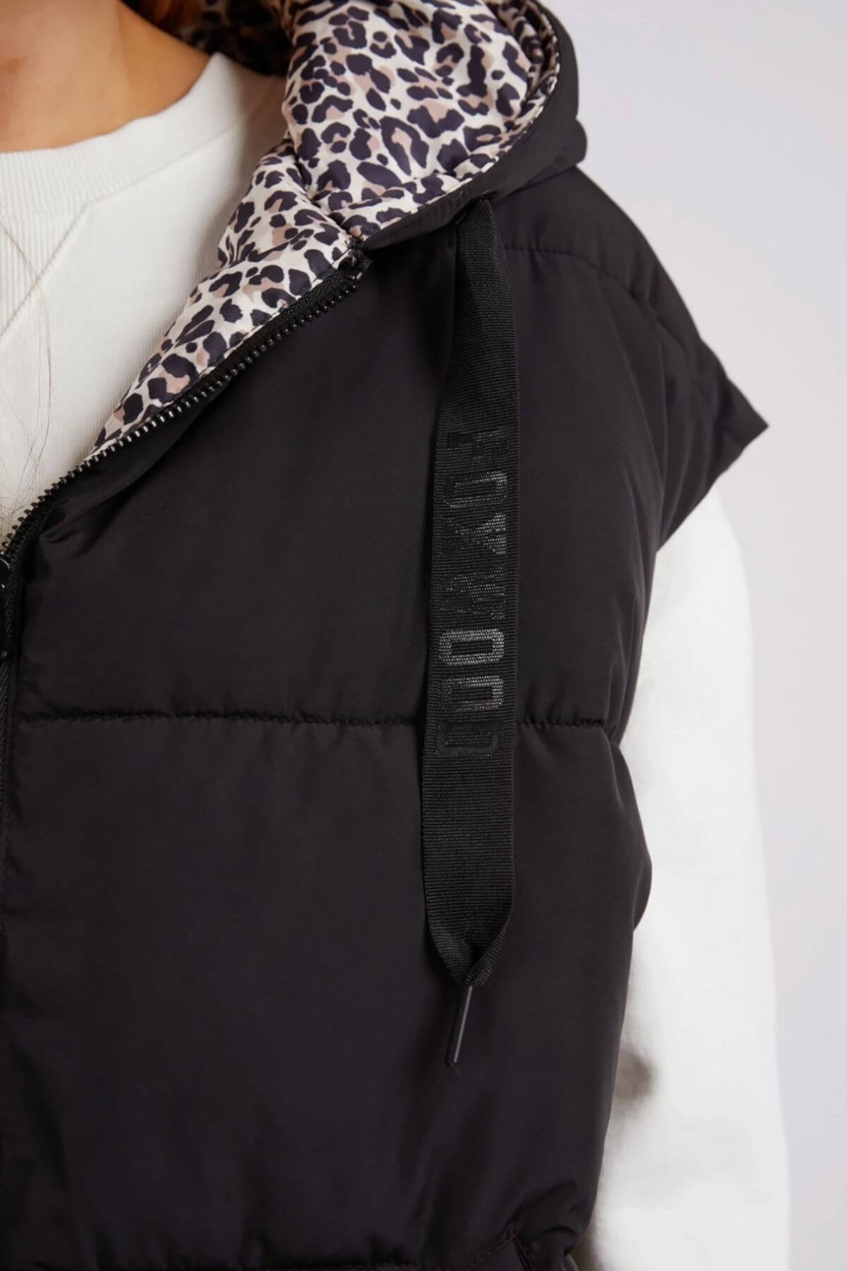 A close-up shows a person wearing the Foxwood Sports Leopard Vest, a reversible piece with a stylish leopard print and adjustable toggles on the black drawstring hood. The vest is paired with a white top, offering flair and functionality.