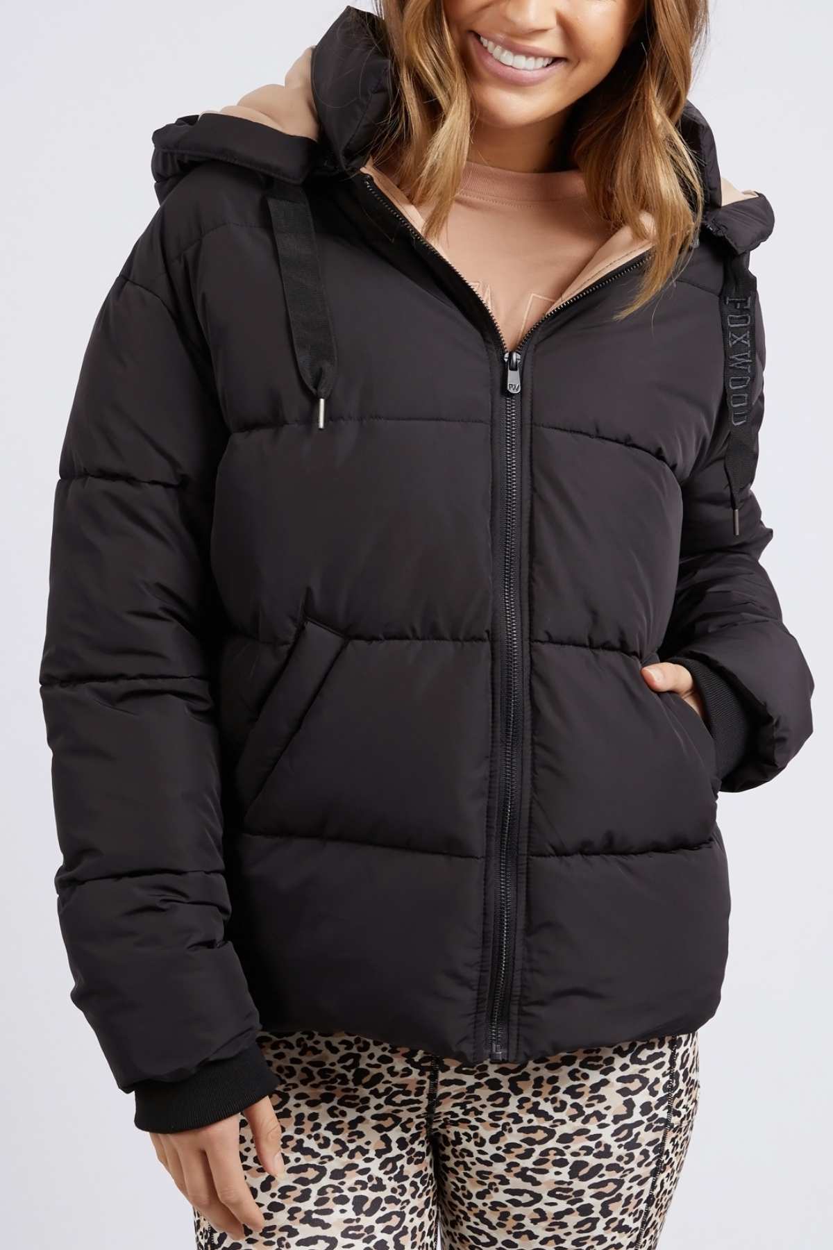 Wearing a black Foxwood Sporty Puffa with an oversized hood over a beige top, a person smiles, placing one hand in the jacket pocket. They pair it with leopard print pants against a plain white background.