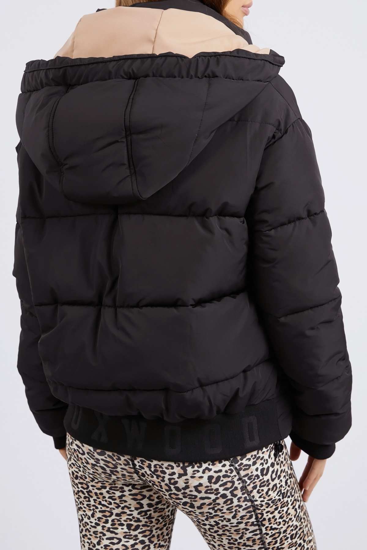Person in a black Foxwood Sporty Puffa jacket with an oversized beige hood faces away, highlighting visible stitching and a ribbed hem with "OXWOOD" text. Paired with leopard print leggings, their outfit showcases versatile flair.