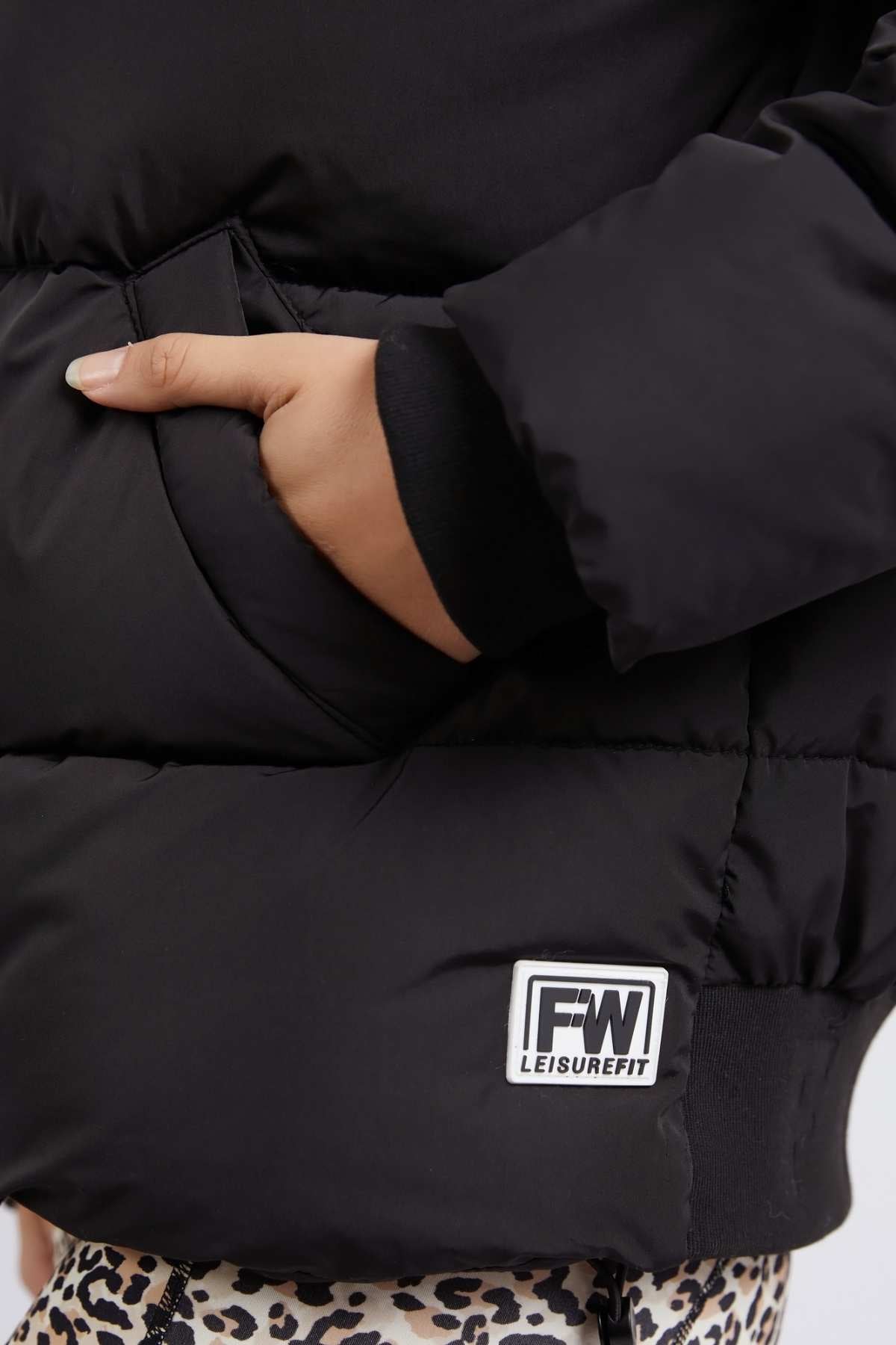 A person wearing a black Foxwood Sporty Puffa jacket with an oversized hood has their hand in the side pocket. The versatile jacket, labeled "FW LEISUREFIT" above the hem, pairs perfectly with their leopard-print pants.
