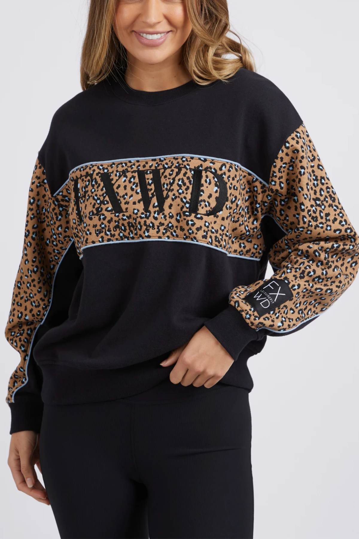 A person wearing the Foxwood The Panel Crew, which is a stylish black sweatshirt featuring "AIVD" embroidered on the chest and bold animal print panels adorning the sleeves. Their wavy, shoulder-length hair frames their face as they exude confidence, posing with one hand resting on their hip.