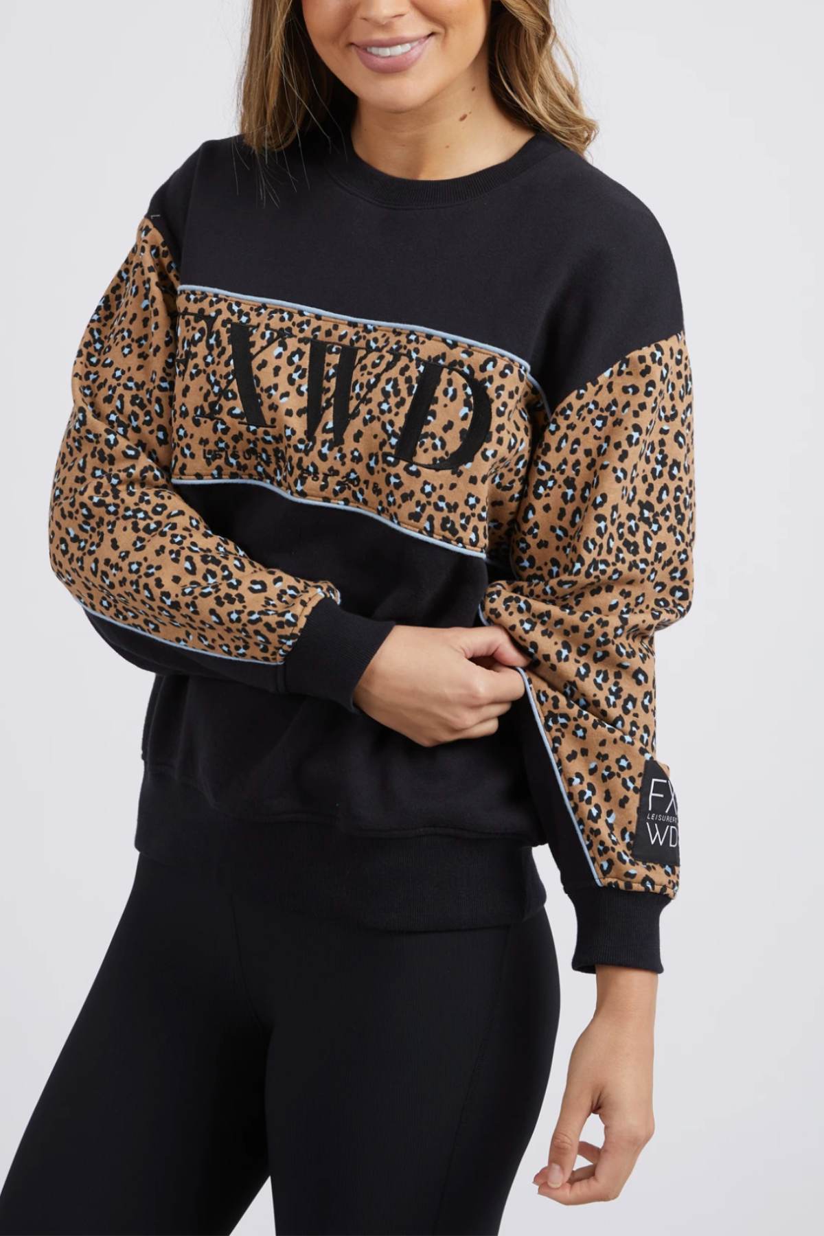 A woman with long hair, wearing the Foxwood The Panel Crew sweatshirt featuring animal print panels and chest embroidery, is standing against a plain background. She has a slight smile as she holds the sleeve of her sweatshirt and pairs it with black leggings.