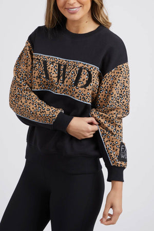 A woman with long hair, wearing the Foxwood The Panel Crew sweatshirt featuring animal print panels and chest embroidery, is standing against a plain background. She has a slight smile as she holds the sleeve of her sweatshirt and pairs it with black leggings.
