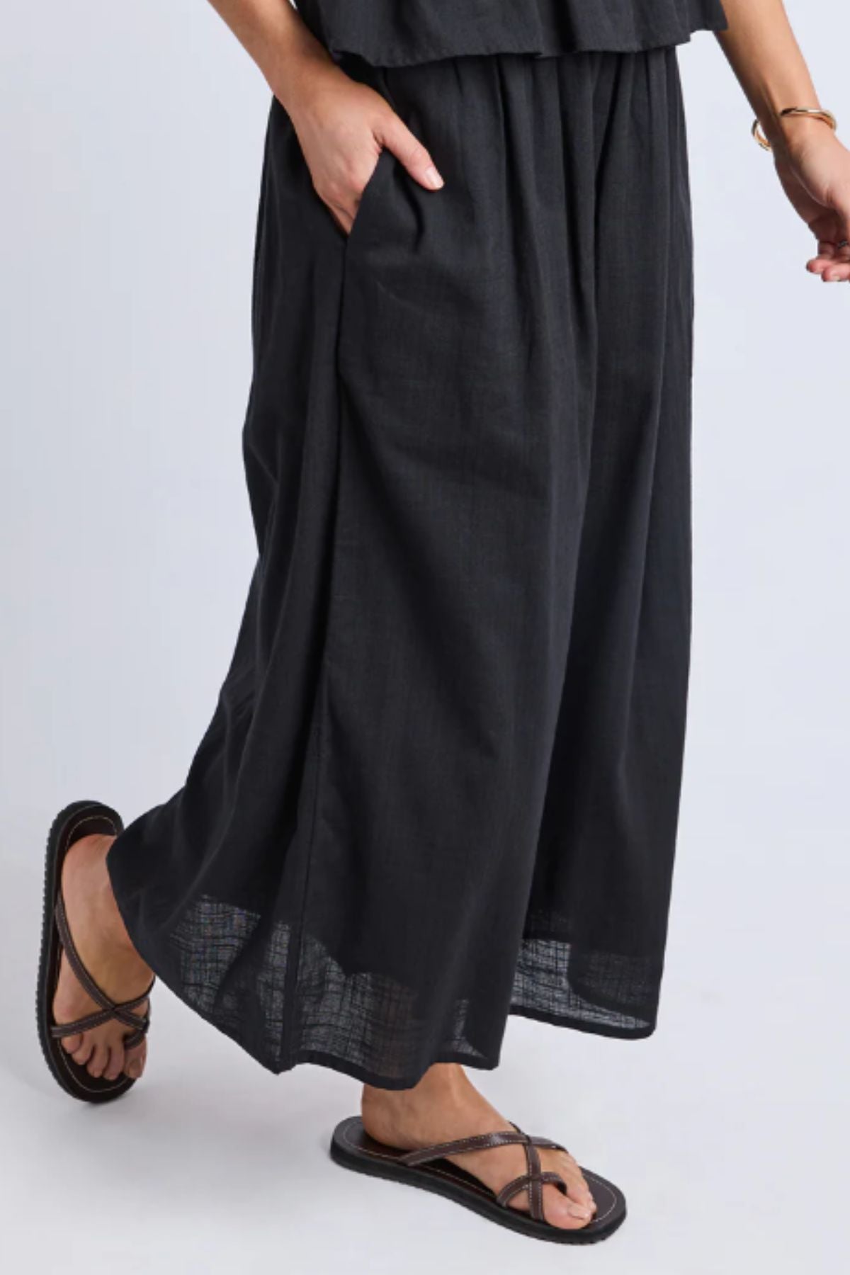 Wearing the Foxwood Tiana Skirt—a loose, ankle-length black maxi with an elastic waist—paired with black strappy sandals and a gold bracelet, one hand in pocket, the person stands against a light gray background.
