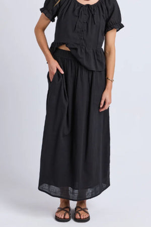 Wearing a black short-sleeve loose blouse, Foxwood Tiana Skirt with elastic waist, and sandals, the outfit is casual and comfortable. The person stands against a plain light background.
