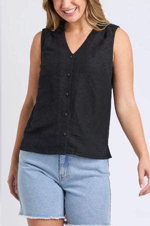 A woman stands smiling with one arm slightly outstretched, wearing the Foxwood Anara Top and light blue denim shorts against a plain background.