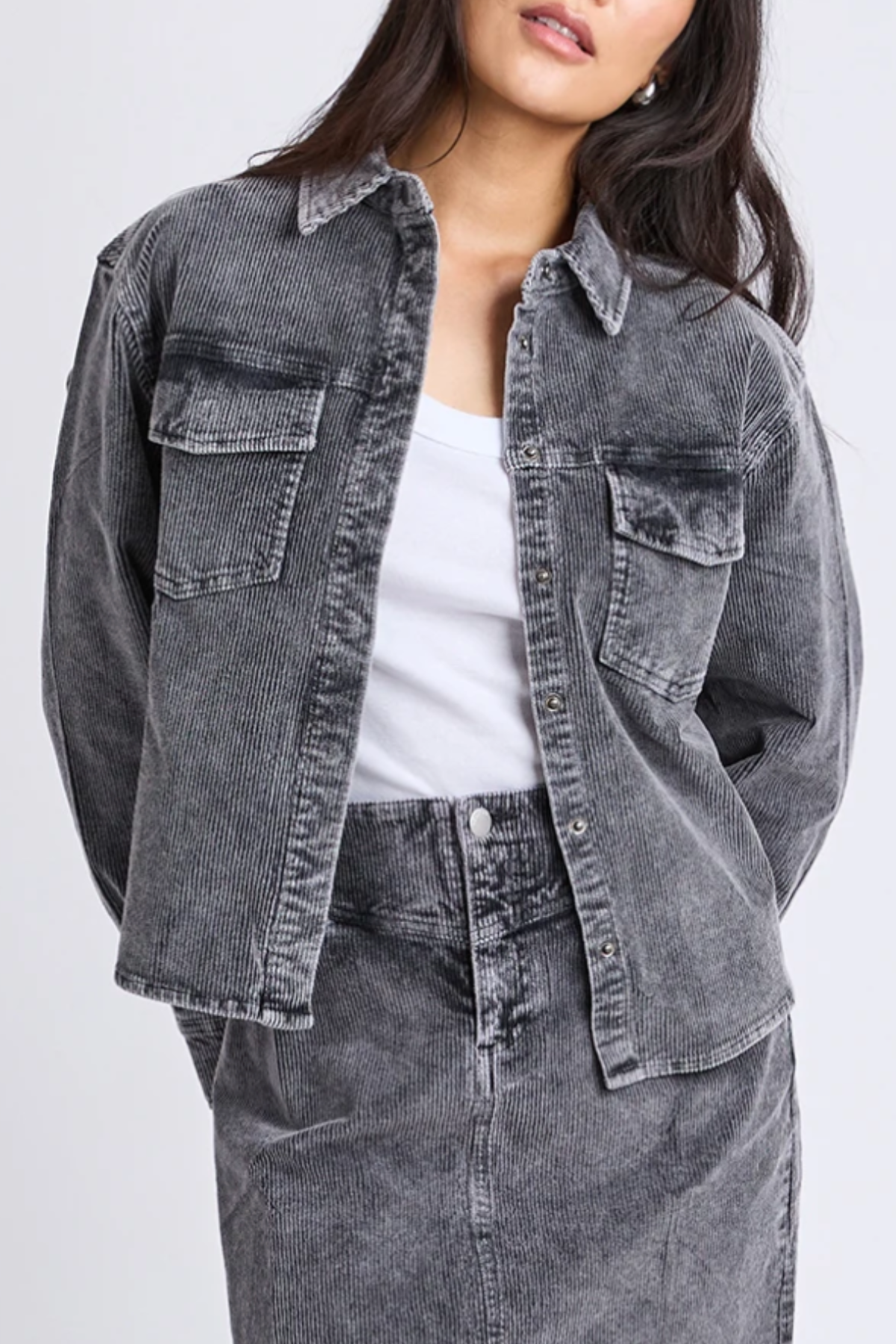 A woman with long dark hair stands against a plain background wearing the Foxwood Tularose Cord Shirt as a grey corduroy jacket with front pockets, styled over a white top and matching skirt, showcasing effortless style.