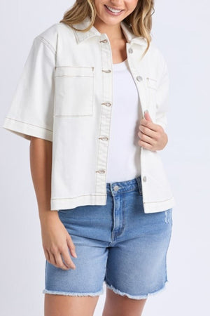 A woman smiles while wearing the Foxwood Valletta Short Sleeve Shirt over a white top with blue denim shorts, holding the shirt open and standing against a plain light background.