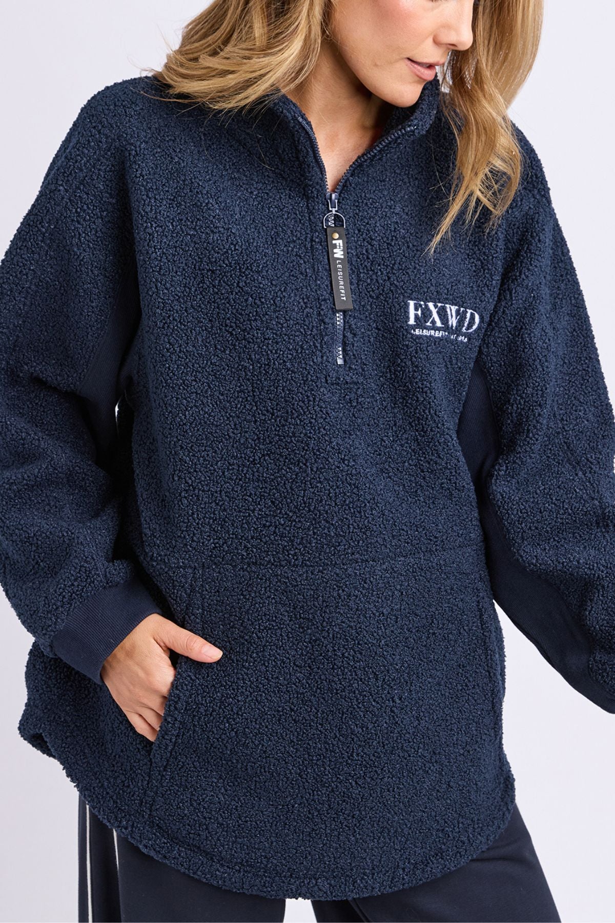 A woman wears the Foxwood Weekend Boucle Crew, a navy blue 1/4 zip sherpa pullover with "FXWD" and small chest text embroidery, her hand in her pocket and gaze down, showcasing cozy loungewear style.