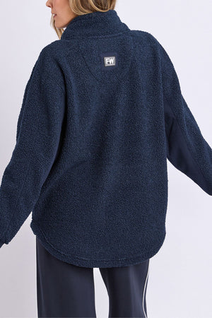 A person with wavy, light brown hair is seen from behind wearing the Foxwood Weekend Boucle Crew, a navy blue fleece with a textured, fluffy look and a rectangular logo patch at the top center.