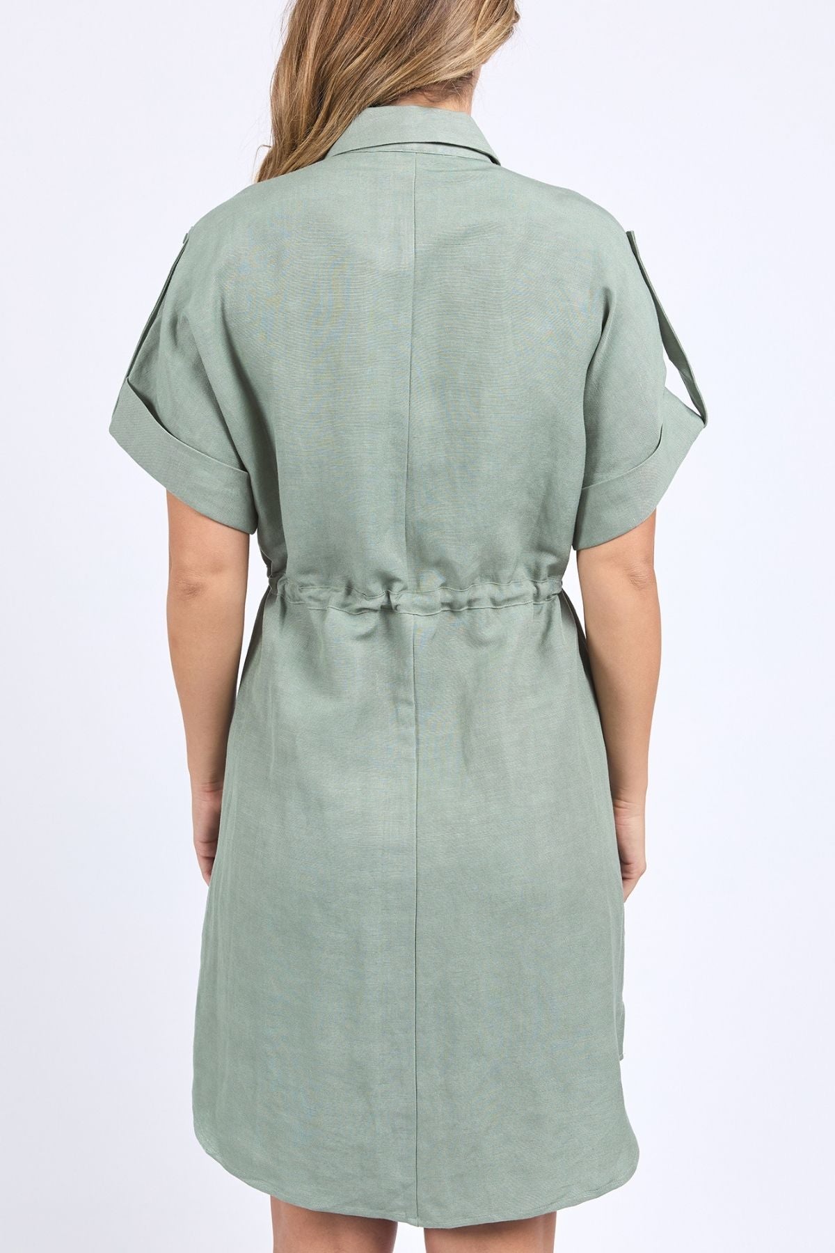 A woman with loose light brown hair faces away, wearing the Foxwood Willow Shirt Dress. This sage green linen-viscose blend dress has a drawstring waist and folded cuffs, set against a plain white background.