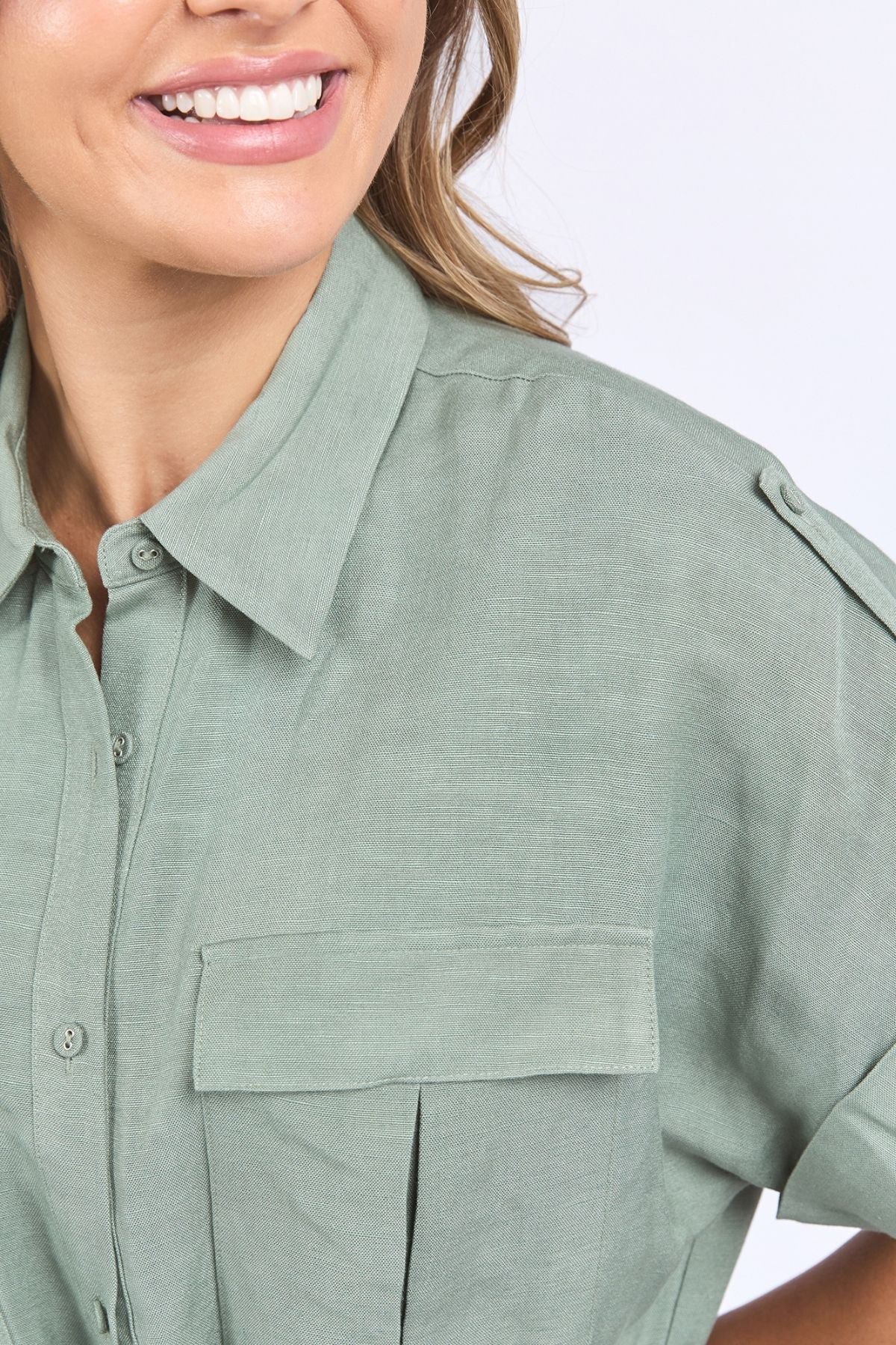 A woman smiles, wearing the Foxwood Willow Shirt Dress in light green linen-viscose, featuring a front pocket and shoulder epaulets, shown from the shoulders up against a light background.
