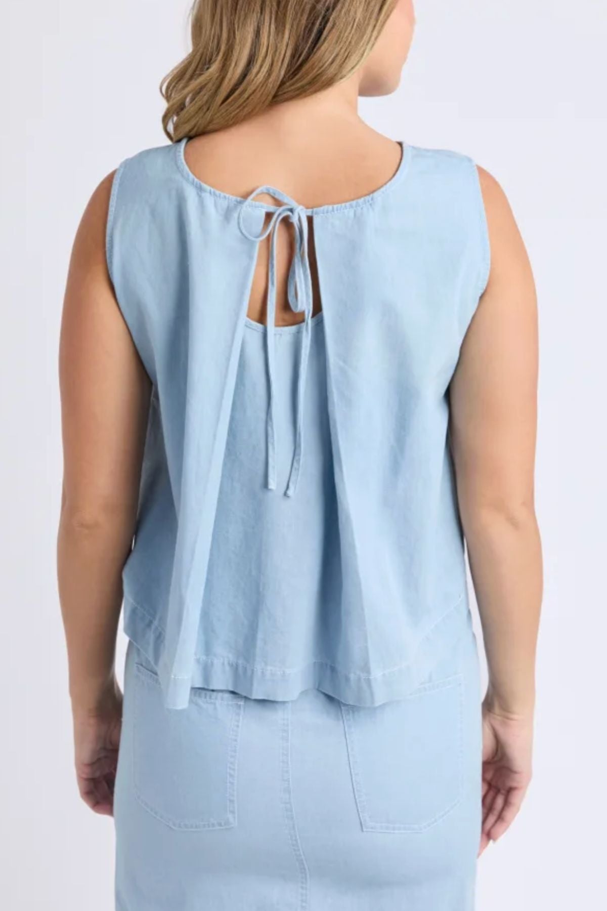 A woman is seen from behind in a Foxwood Zetta Top, a chambray sleeveless style with a back tie, paired with a matching light blue skirt; her light brown hair falls over one shoulder for an effortless summer look.