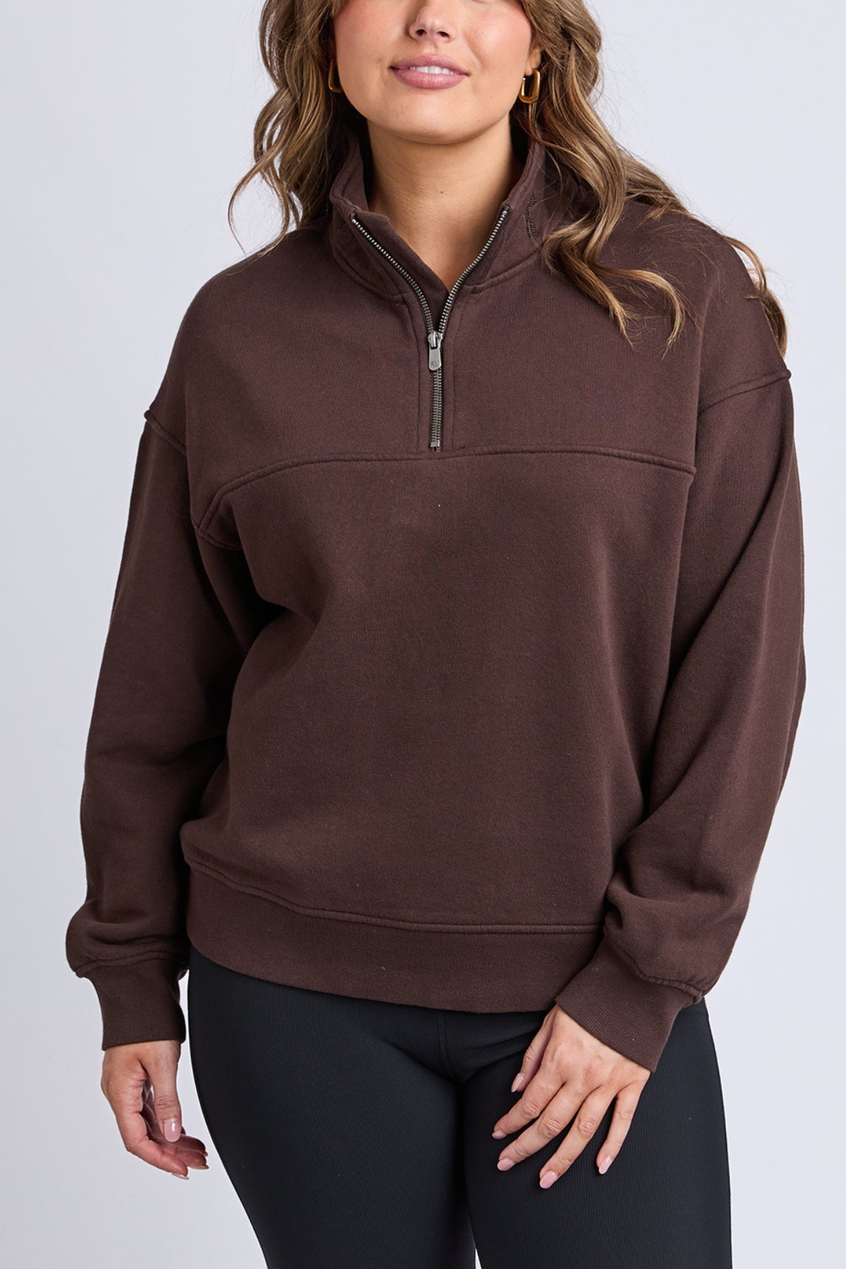 A woman wearing the Foxwood College Zip Crew in dark brown with black leggings stands against a plain light background, her wavy hair framing her face as she rests one hand on her hip.