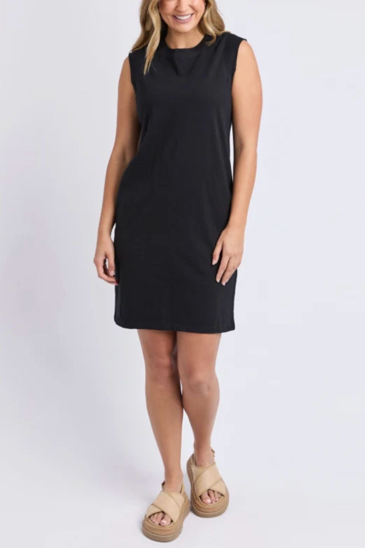 A woman smiles against a plain background in the knee-length, black Foxwood Reform Dress with a relaxed fit, styled with beige platform sandals and her wavy shoulder-length hair.