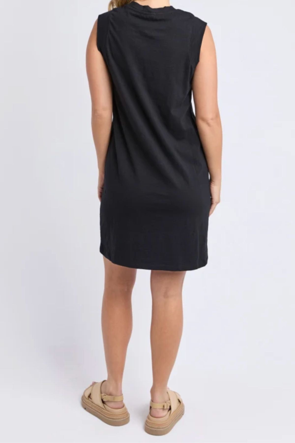 A person stands facing away, wearing the Foxwood Reform Dress—a relaxed-fit, sleeveless black cotton dress—paired with beige platform sandals, set against a plain light background.