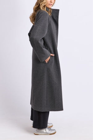 A woman in profile stands against a light background, wearing the Foxwood Fermont Coat over black pants and silver sneakers. With one hand in her pocket and loose wavy hair, she models an ideal transitional look.