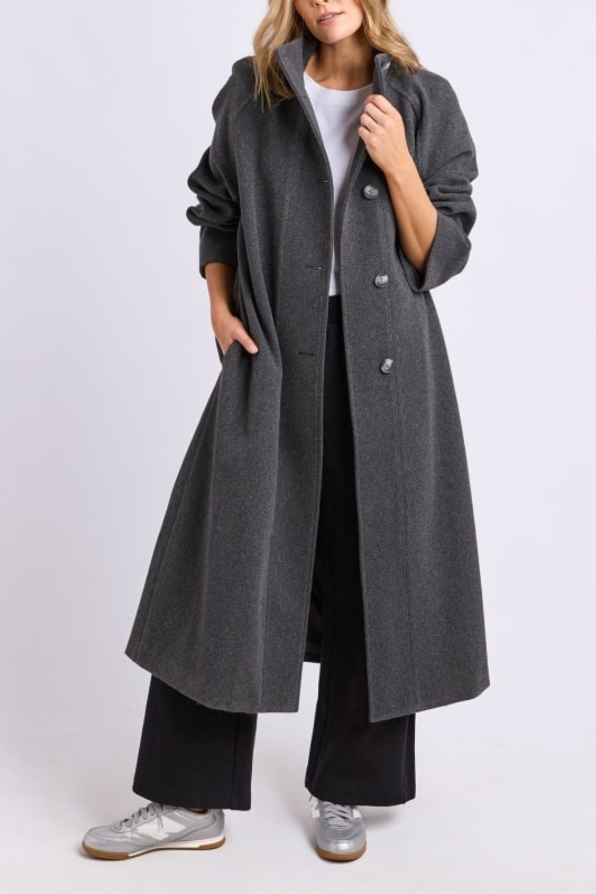 A woman wears the Foxwood Fermont Coat in dark gray over a white shirt and black wide-leg pants, paired with light gray sneakers. Her hands rest in her coat pockets and her loose wavy hair frames her face.