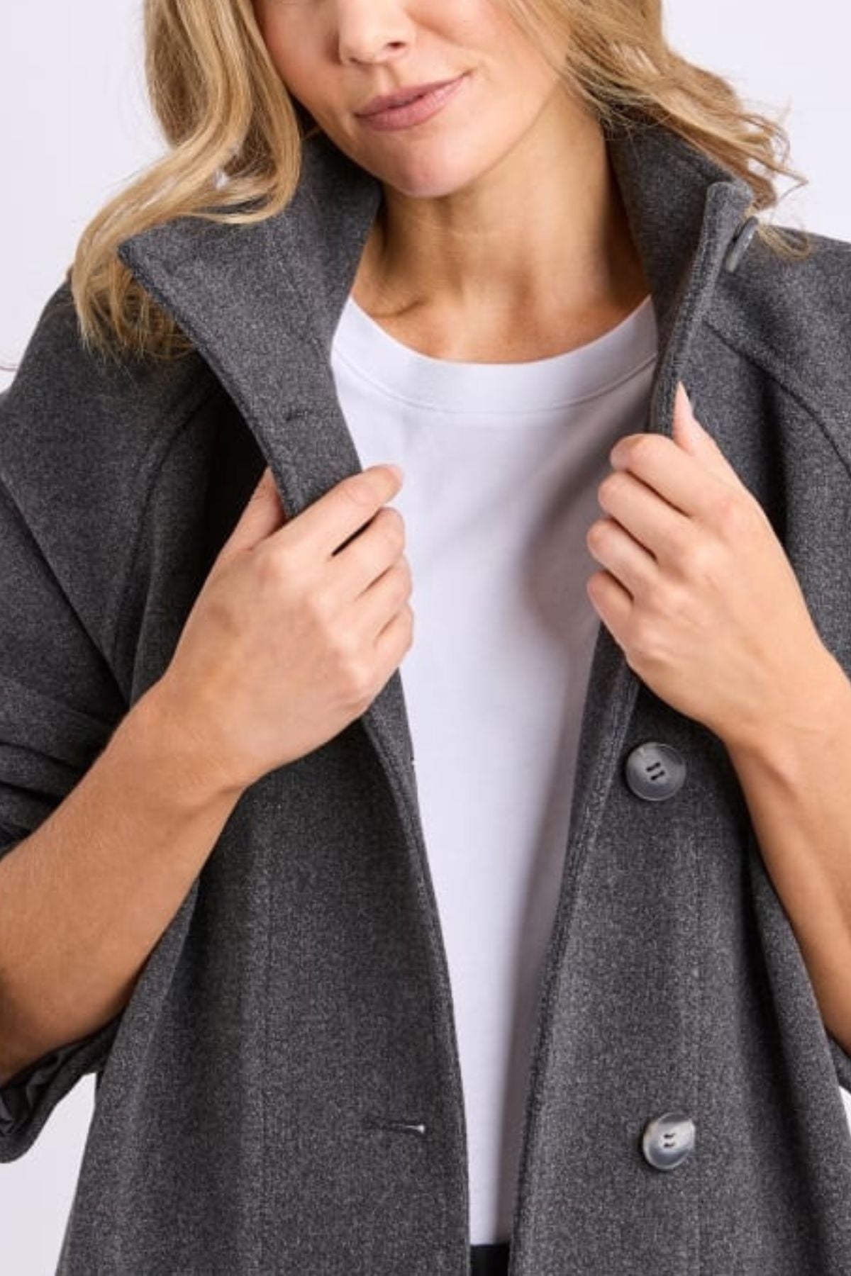A woman in a white t-shirt and the Foxwood Fermont Coat, a stylish jacket with large buttons, stands facing the camera while holding the open collar. Her face is partially visible.