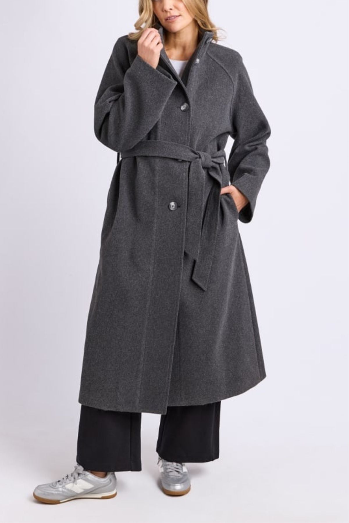 A person models the Foxwood Fermont Coat—a long women's coat—in dark gray, worn over wide-leg black pants and silver sneakers, standing with one hand in the pocket against a plain light background.