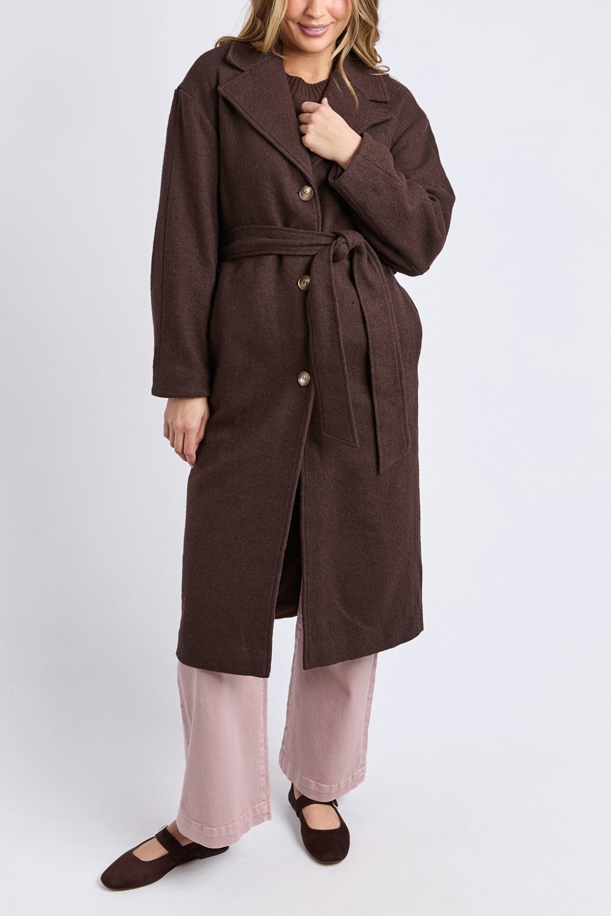 A model wears the Foxwood Whistler Coat—long, brown, with a belt—crafted from a wool-polyester blend, over light pink pants and brown shoes. The women's coat is buttoned and tied at the waist, set against a plain white background.