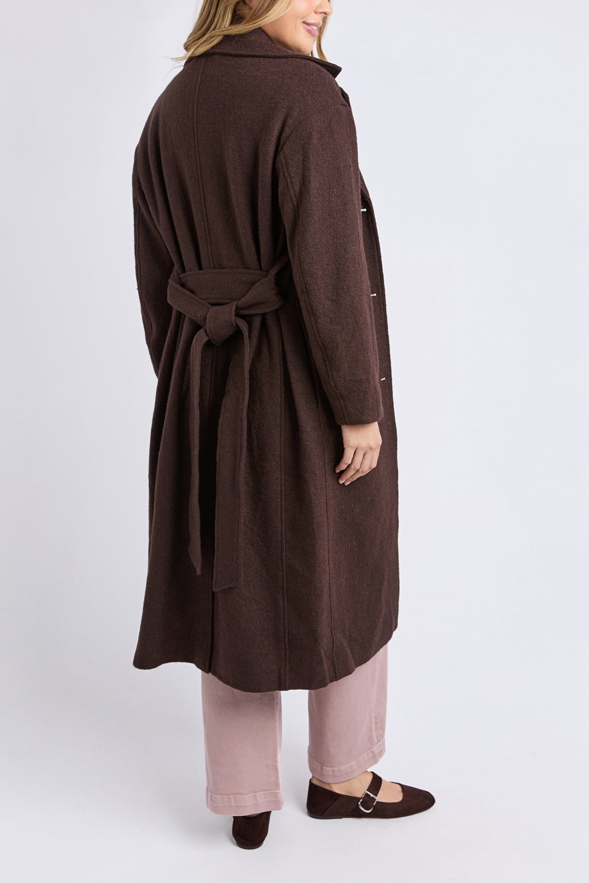 Woman standing and smiling, shown from the back against a plain light background, wearing the Foxwood Whistler Coat—a stylish brown wool-polyester blend women’s coat with a tied back belt—paired with pink pants and brown shoes.